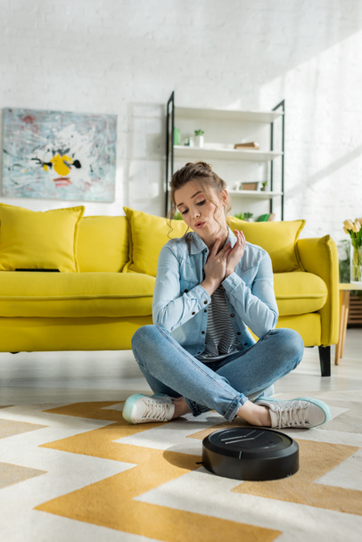 young woman looking at robotic vacuum cleaner on carpet in living room  - Valokuva, kuva