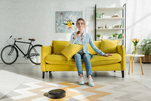 attractive woman watching tv while robotic vacuum cleaner washing carpet in living room  - Foto, afbeelding