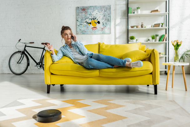 attractive girl talking on smartphone while robotic vacuum cleaner washing carpet in living room  - Foto, afbeelding