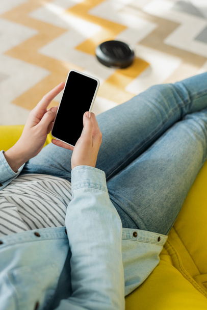 cropped view of woman holding smartphone with blank screen while robotic vacuum cleaner washing carpet in living room  - Foto, afbeelding