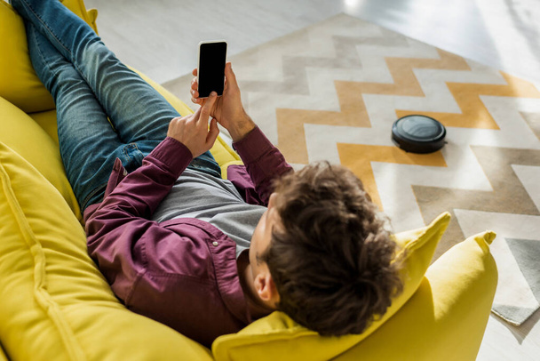 overhead view of man relaxing on sofa and holding smartphone with blank screen while robotic vacuum cleaner washing carpet in living room - Foto, imagen