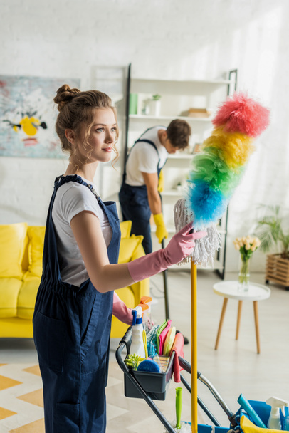 selective focus of attractive cleaner holding duster brush near cleaning trolley - Foto, imagen