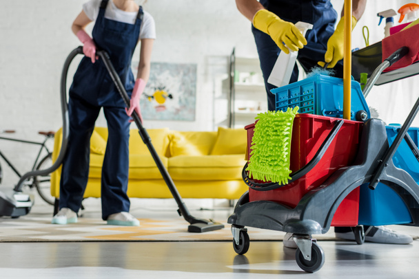 cropped view of cleaner in rubber gloves holding spray bottle near cleaning trolley and coworker with vacuum cleaner  - Photo, Image