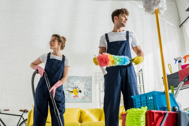 selectieve focus van happy cleaners in uniform weg te kijken in de buurt van schoonmaakwagen in de moderne woonkamer  - Foto, afbeelding