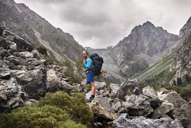 Portrait of tourist with big backpack is on the rocky trail in the mountain valley of Karakol national park, Kyrgyzstan - Zdjęcie, obraz