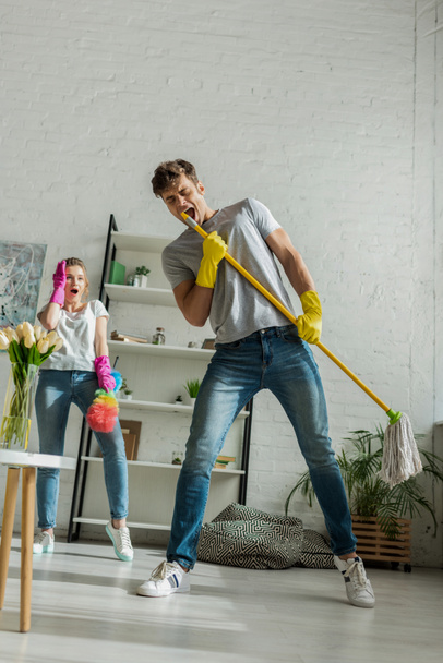 young and surprised woman looking at handsome man singing while holding mop  - Photo, Image