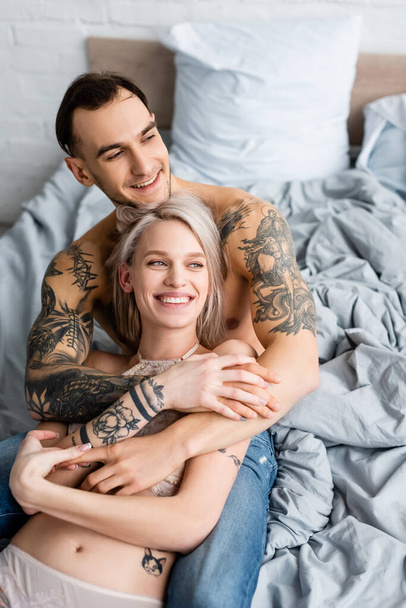 Smiling shirtless man hugging tattooed girlfriend in lingerie on bed - Foto, Imagen