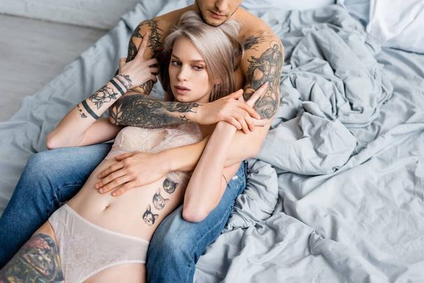 М'язистий чоловік в джинсах обіймає татуйовану жінку в нижній білизні на ліжку
  - Фото, зображення