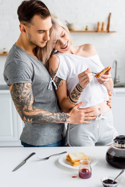 Handsome tattooed man hugging smiling girlfriend with toast near coffee pot and jams on kitchen table - Fotoğraf, Görsel