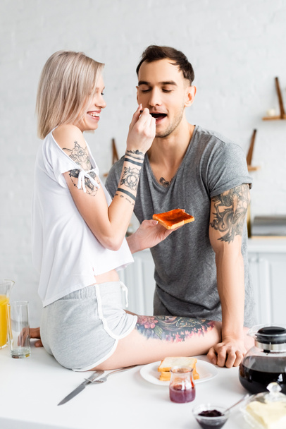 Smiling woman sitting on kitchen table and feeding tattooed boyfriend with jam during breakfast in kitchen  - Фото, изображение