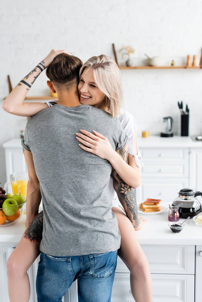 Mujer sonriente abrazando novio tatuado cerca del desayuno en la mesa de la cocina
  - Foto, Imagen