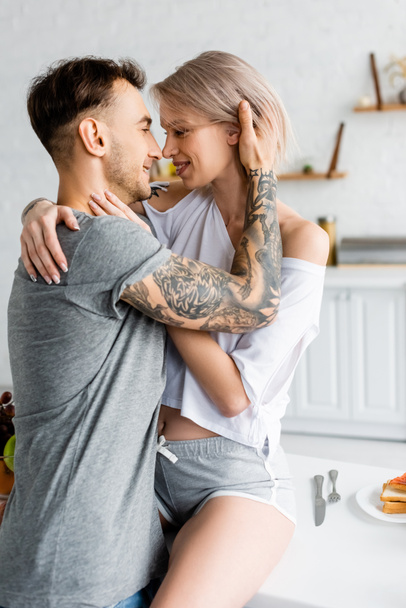 Vista lateral del hombre tatuado abrazando a su novia sonriente en la mesa de la cocina
  - Foto, Imagen