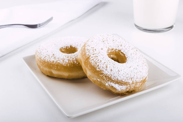 Donuts covered with icing sugar on a plate on a white background accompanied by a glass of milk and a fork on a napkin. Delicious breakfast or lunch. American food. Bakery and pastry products. - Photo, Image