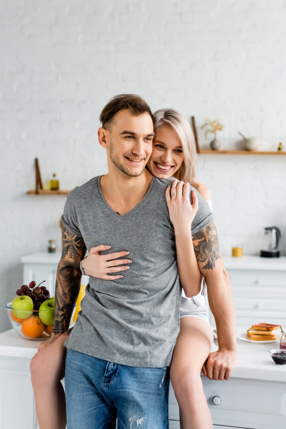 Привлекательная женщина обнимает улыбающегося парня с татуировкой рядом с тостами и фруктами на кухонном столе
  - Фото, изображение