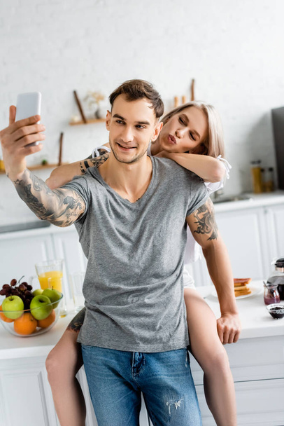 Selective focus of woman grimacing while taking selfie near tattooed boyfriend in kitchen  - Fotoğraf, Görsel