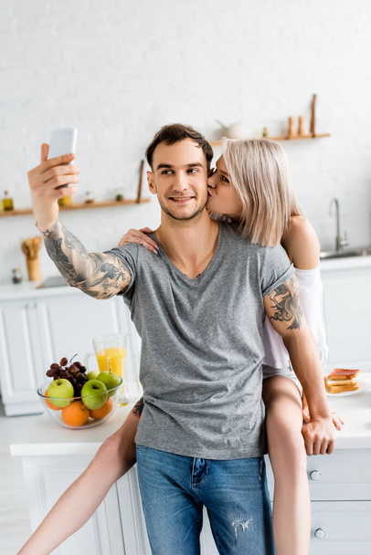 Woman kissing tattooed boyfriend taking selfie with smartphone in kitchen  - Fotoğraf, Görsel