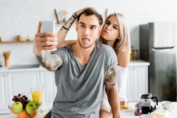 Selective focus of tattooed couple grimacing while taking selfie with smartphone during breakfast in kitchen  - Fotografie, Obrázek