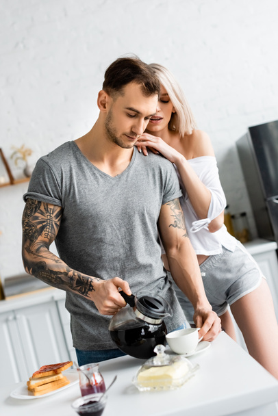 Attrayant femme étreignant beau petit ami tatoué avec cafetière près du petit déjeuner sur la table de cuisine
  - Photo, image