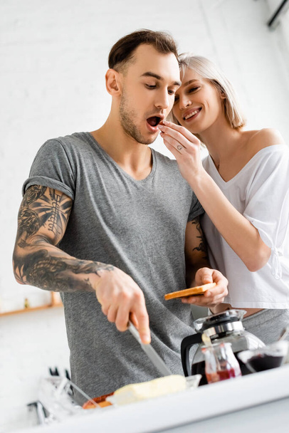 Enfoque selectivo de la mujer sonriente alimentando novio tatuado con uva durante el desayuno en la cocina
  - Foto, Imagen