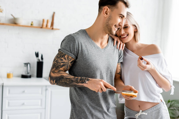 Beautiful smiling woman holding grape and embracing tattooed boyfriend with toast in kitchen  - Photo, Image