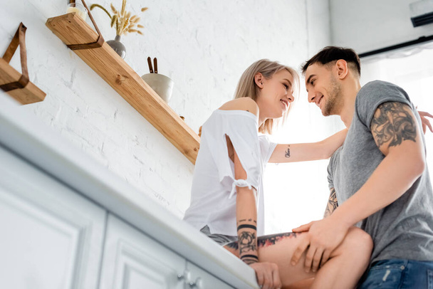 Low angle view of handsome man smiling at tattooed girlfriend on kitchen worktop  - Photo, Image