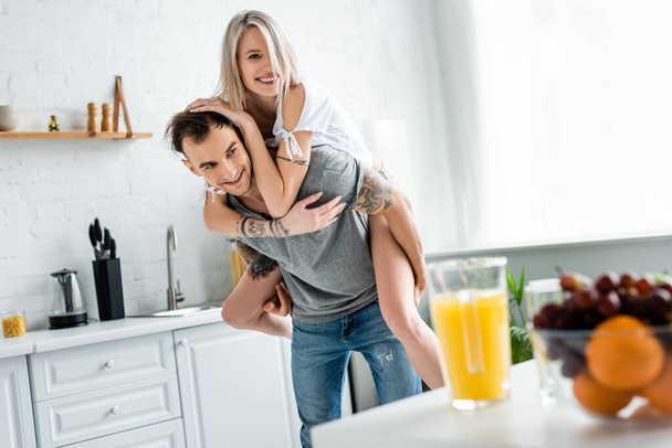 Selective focus of smiling girl piggybacking on tattooed boyfriend near fruits and orange juice on kitchen table  - Foto, Imagen
