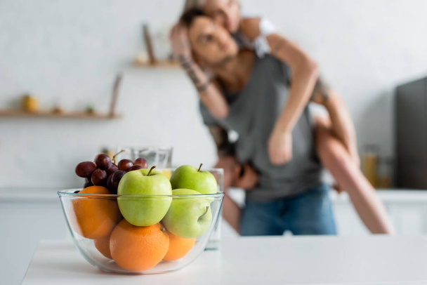 Selective focus of fresh fruits in bowl on table and woman piggybacking on boyfriend in kitchen   - 写真・画像