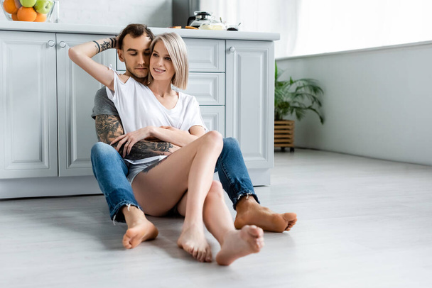 Smiling girl embracing tattooed boyfriend on floor in kitchen  - Φωτογραφία, εικόνα