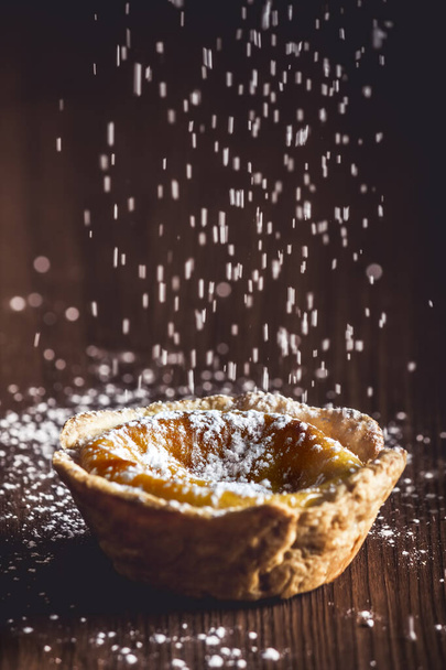 Artisan cake from Belem, traditional dessert from Portugal. Delicious handmade Belem pie that is being sprinkled with icing sugar on a rustic dark wood table. Cream cake. - Fotó, kép