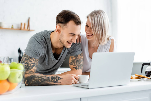Mujer sonriente abrazando novio tatuado cerca de la computadora portátil y café en la mesa de la cocina
 - Foto, imagen