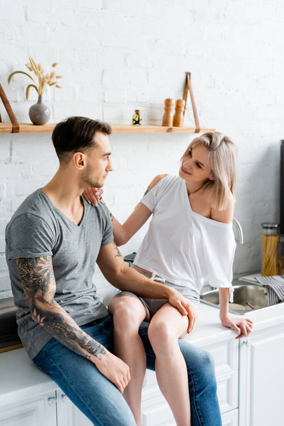 Tattooed man touching leg of smiling girlfriend on kitchen worktop  - Fotó, kép
