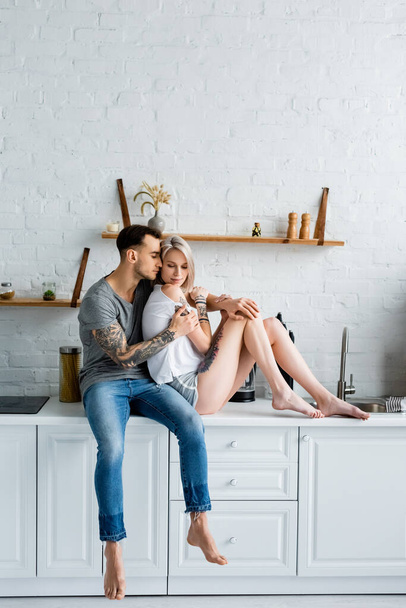 Tattooed man hugging attractive girlfriend while sitting on kitchen worktop  - 写真・画像