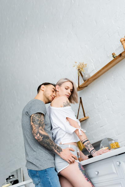 Χαμηλή γωνία άποψη του όμορφου άντρα με τατουάζ φιλώντας το λαιμό της ξανθιάς κοπέλας στην κουζίνα  - Φωτογραφία, εικόνα