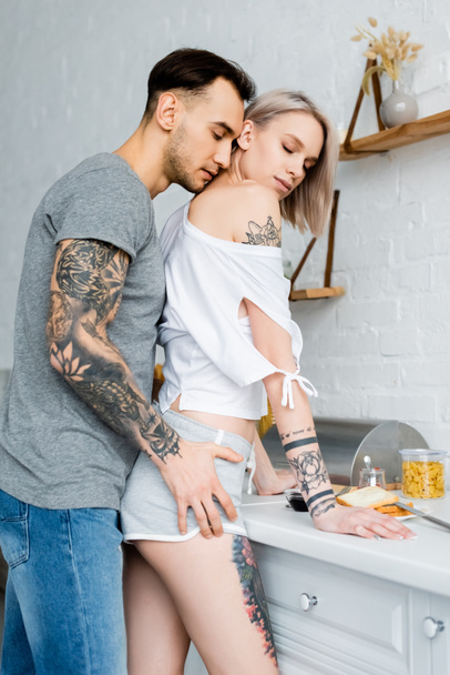 Zijaanzicht van getatoeëerde man aanraken heup van mooie vriendin in de buurt van het ontbijt op keuken werkblad  - Foto, afbeelding
