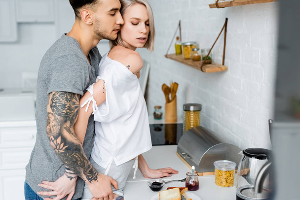 Side view of tattooed man touching beautiful blonde girlfriend near breakfast in kitchen  - Photo, Image