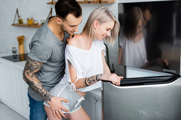 Side view of tattooed man touching beautiful smiling girlfriend near fridge in kitchen  - Fotografie, Obrázek