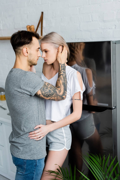 Oldalra néző jóképű férfi megérinti gyönyörű tetovált barátnő közelében hűtőszekrény a konyhában  - Fotó, kép