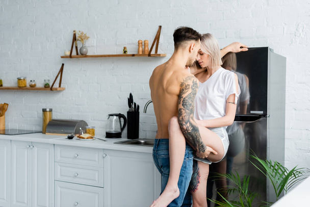 Shirtless tattooed man touching leg of beautiful girlfriend near fridge in kitchen  - Photo, Image