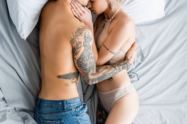 Top view of muscular tattooed man hugging woman in underwear on bed  - Fotografie, Obrázek