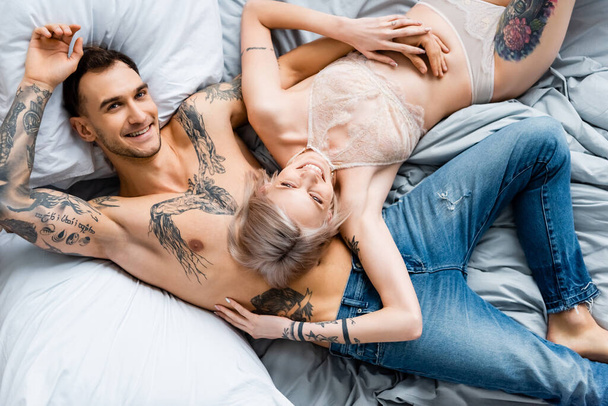 Top view of tattooed man smiling at camera near girlfriend in lingerie on bed  - 写真・画像