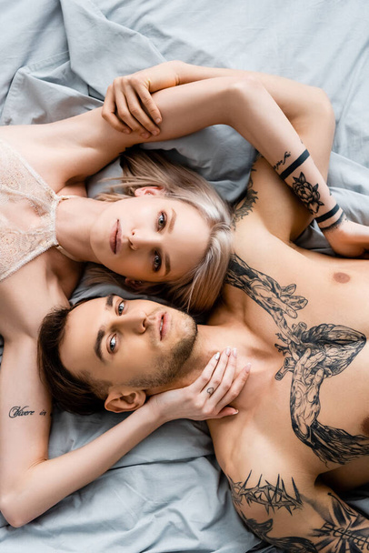 Blick von oben auf tätowiertes Paar, das in die Kamera schaut, während es sich auf dem Bett berührt  - Foto, Bild
