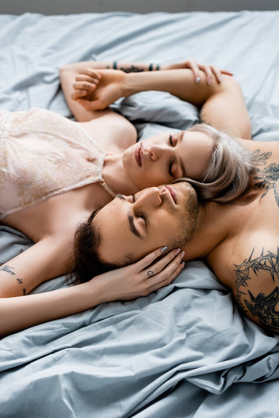 Άνδρας χωρίς πουκάμισο με τατουάζ ξαπλωμένος δίπλα σε μια κοπέλα με σουτιέν στο κρεβάτι απομονωμένος στο γκρι - Φωτογραφία, εικόνα