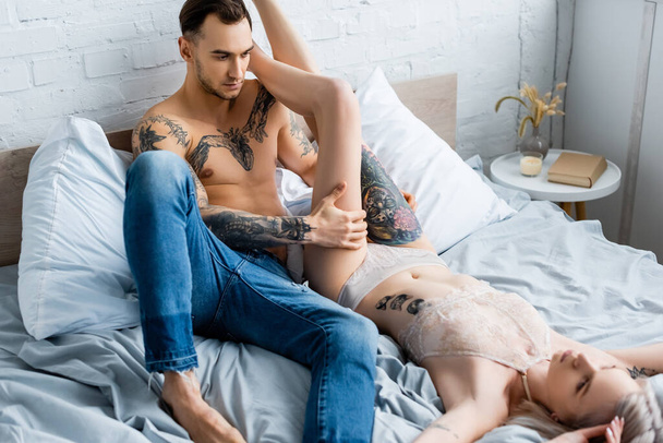 Selective focus of shirtless man looking at sensual tattooed girl in underwear on bed  - Fotó, kép