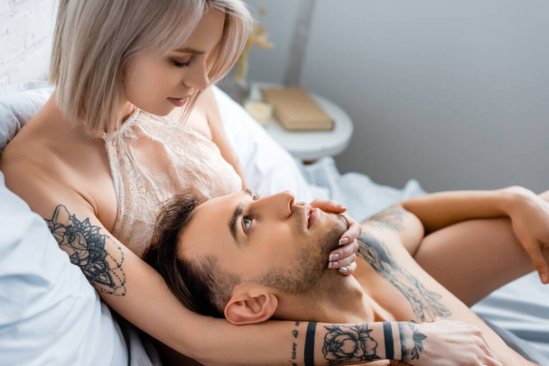 Widok z boku blondynka dziewczyna w biustonosz dotykając tatuaż chłopak na łóżku  - Zdjęcie, obraz