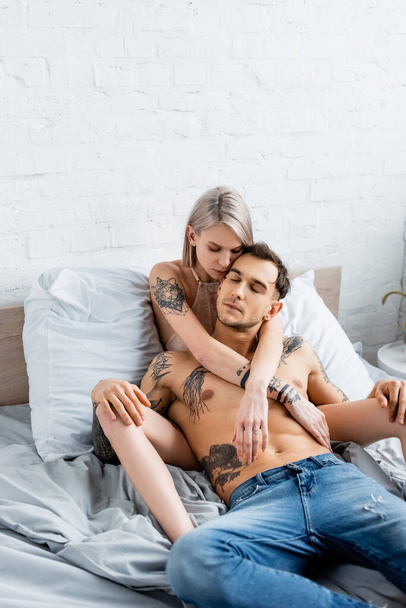 Ξανθιά γυναίκα με σουτιέν που αγγίζει το αγόρι της χωρίς μπλούζα και τατουάζ στο κρεβάτι  - Φωτογραφία, εικόνα