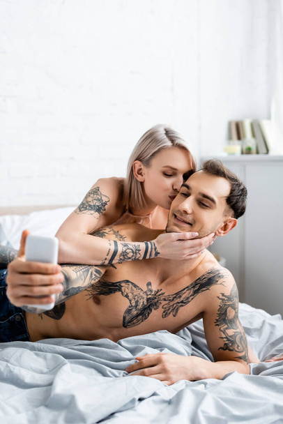 Beautiful girl kissing shirtless tattooed boyfriend taking selfie with smartphone on bed  - Photo, Image