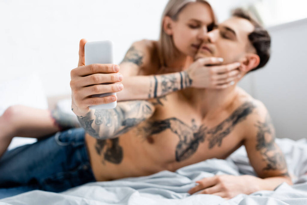 Enfoque selectivo de la chica besando novio tatuado tomando selfie con teléfono inteligente en la cama
   - Foto, Imagen