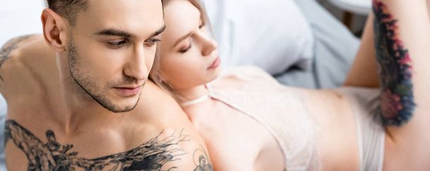 Yatakta seksi kız arkadaşının yanında oturan kaslı dövmeli adamın panoramik fotoğrafı.  - Fotoğraf, Görsel