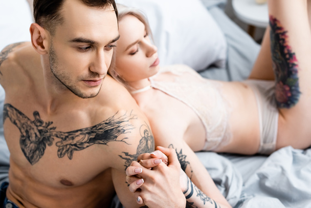 Selective focus of tattooed man holding hand of sensual woman in lingerie on bed  - Фото, изображение