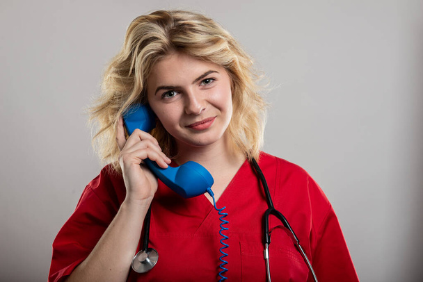 Πορτρέτο του νοσηλευτή φορώντας κόκκινο scrub μιλώντας στο τηλέφωνο στο στούντιο γκρι φόντο - Φωτογραφία, εικόνα